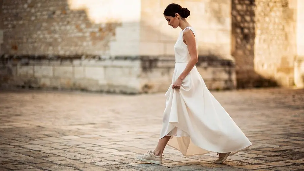 Jeune mariée marchant naturellement dans une cour pavée française, portant une robe de mariée moderne et des baskets beige-ivoire